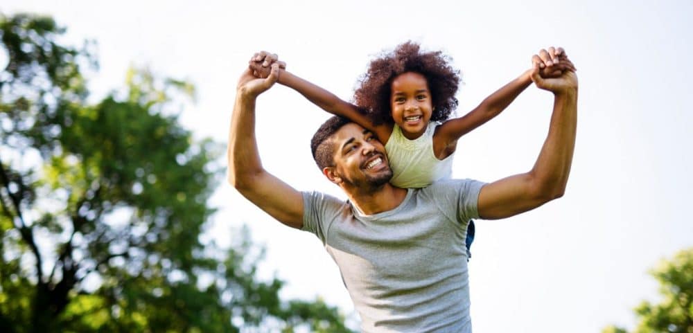 Father carrying his young daughter on his shoulders, both smiling as they hold hands in the air, with a bright sky and green trees in the background peoria az
