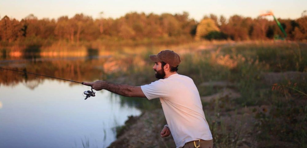 Man in a white shirt and cap fishing at a calm lake, surrounded by grassy fields and trees, casting his line as the sun sets in the background peoria az
