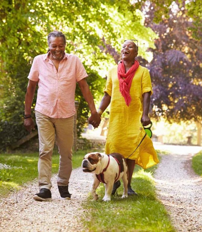 Smiling couple holding hands while walking their bulldog on a tree-lined path, with the woman wearing a yellow dress and red scarf and the man in a pink shirt peoria az