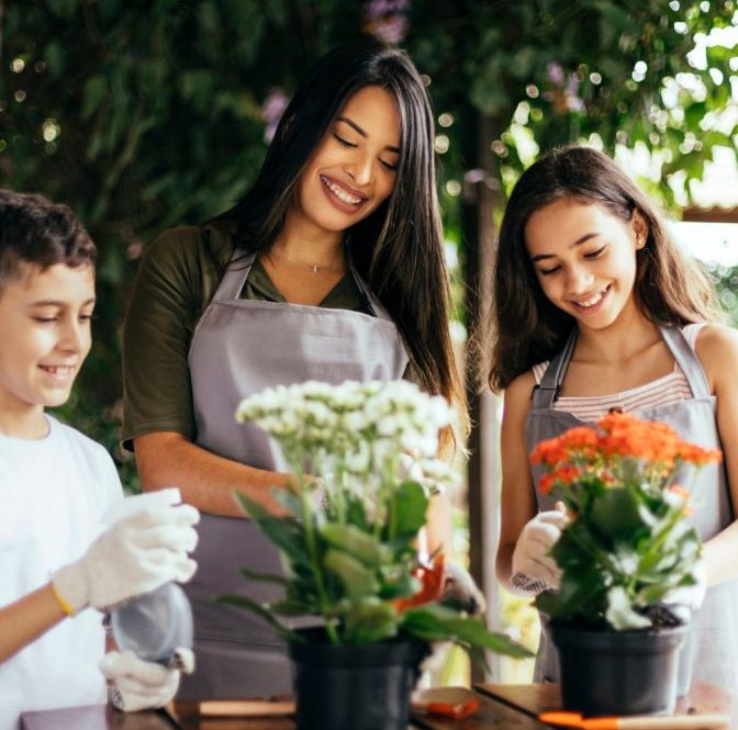 A woman and two children wearing aprons and gardening gloves, smiling while potting flowers in an outdoor setting surrounded by greenery peoria az