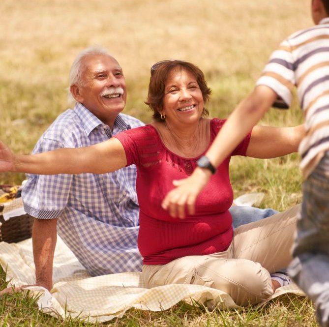 Smiling grandparents sitting on a picnic blanket in a grassy field, with the grandmother reaching out to embrace a running child peoria az
