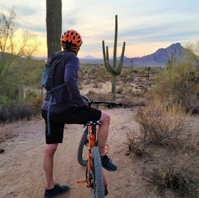 Cyclist wearing an orange helmet and backpack, pausing on a desert trail surrounded by saguaro cacti, with mountains and a sunset in the background peoria az