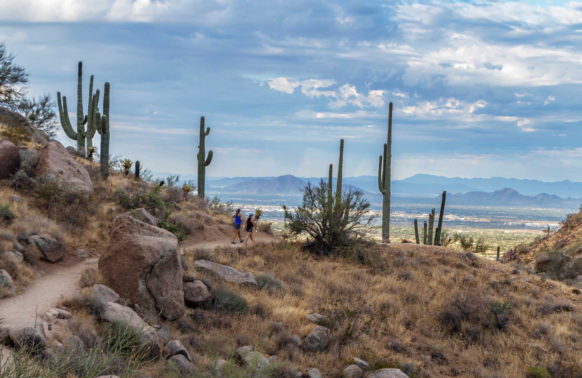 Two hikers walking along a scenic desert trail with large boulders, tall saguaro cacti, and a mountain view in the distance under a partly cloudy sky peoria az