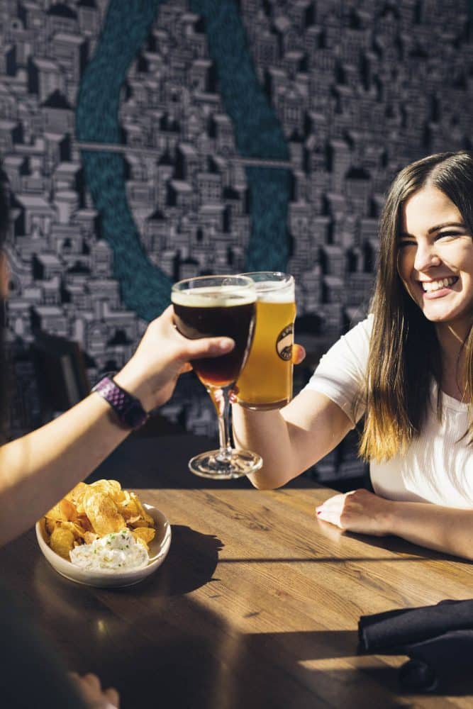 Smiling woman clinking beer glasses with a friend at a bar, with a bowl of chips and dip on the table and a patterned wall in the background peoria az