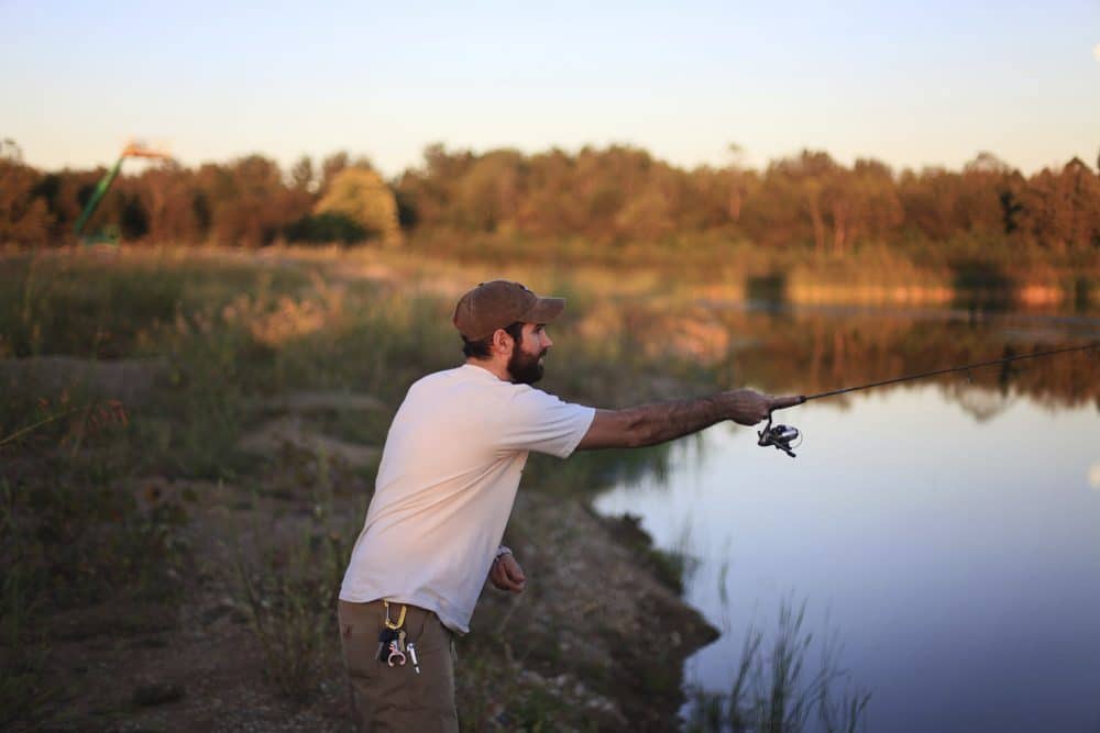 Man in a white shirt and cap fishing at a calm lake, surrounded by grassy fields and trees, casting his line as the sun sets in the background peoria az