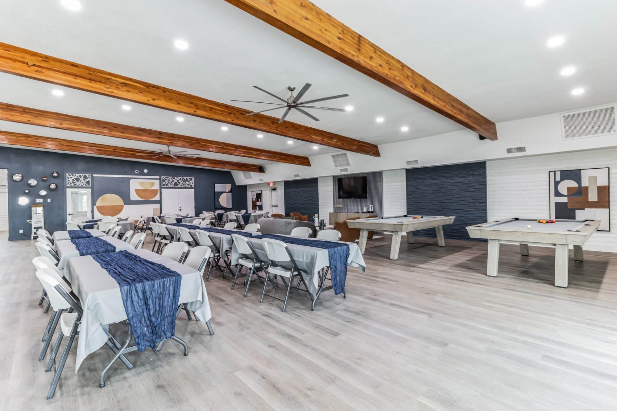 Spacious clubhouse with dining tables, two pool tables, and modern décor, featuring exposed wooden beams, a ceiling fan, and blue accent walls peoria az