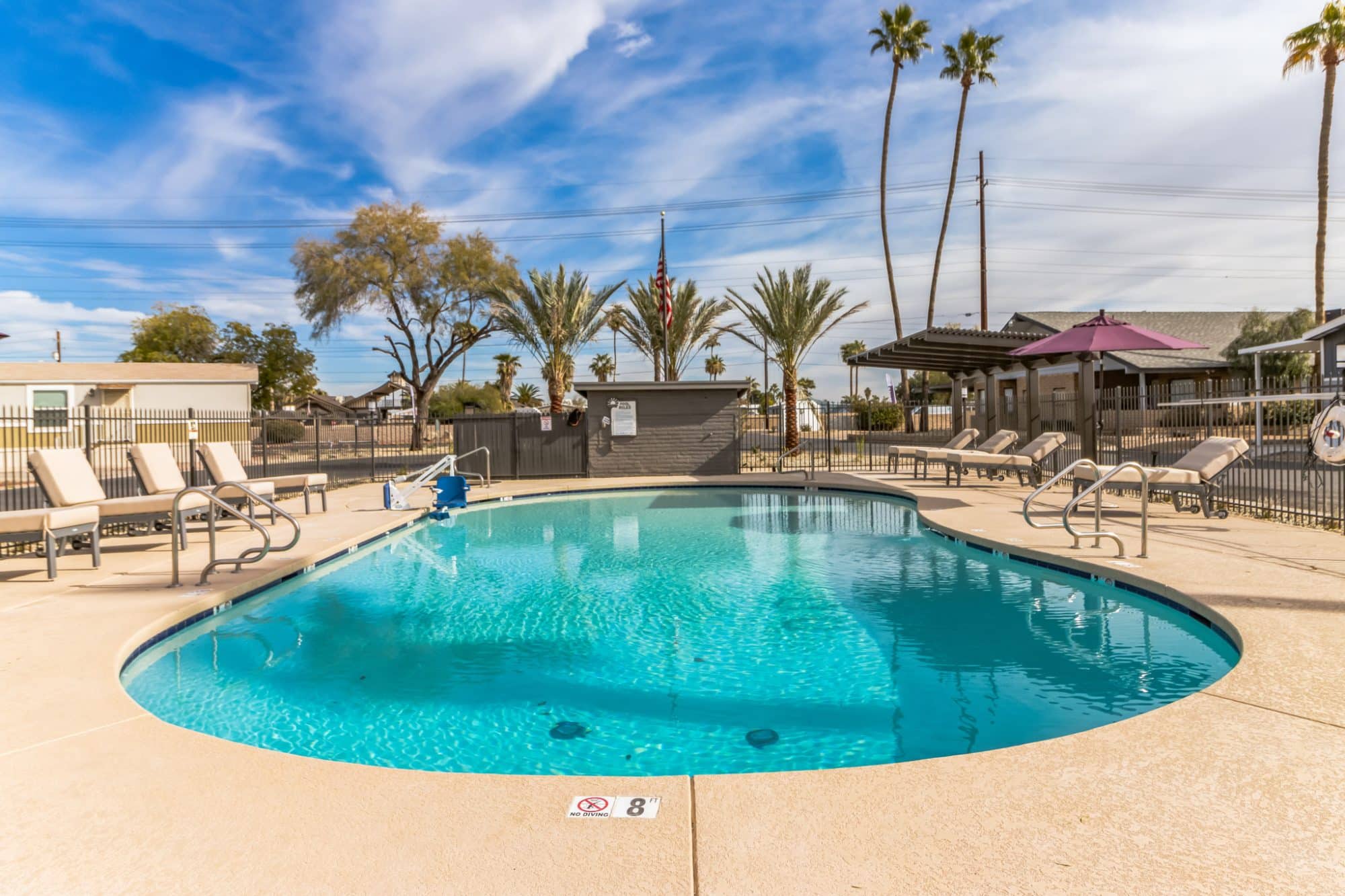 An outdoor swimming pool with clear blue water, surrounded by lounge chairs, palm trees, and a shaded seating area under a bright sky peoria az