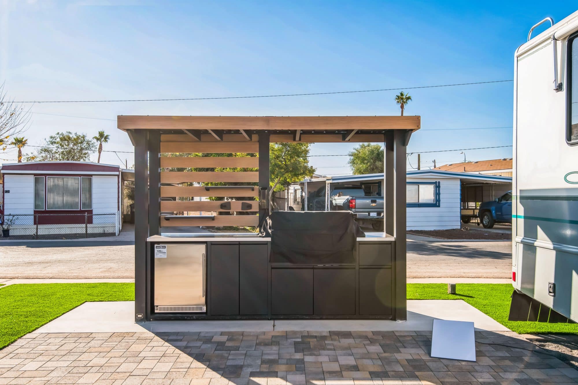Outdoor kitchen with a black countertop, built-in grill, and mini fridge under a pergola, situated in a residential area with parked vehicles in the background peoria az