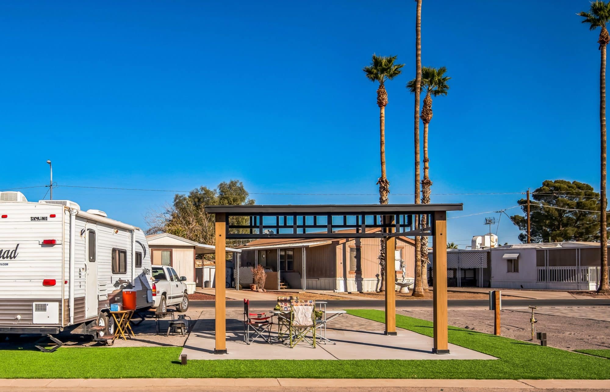 RV site with a white camper, a covered pergola, camping chairs, and a picnic table on artificial grass, surrounded by mobile homes and tall palm trees peoria az