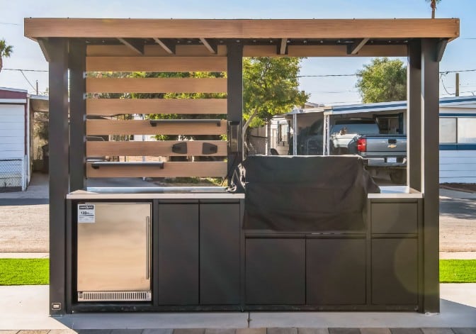 A modern outdoor barbecue station with a covered grill, stainless steel appliances, and black cabinetry, set in a community area with mobile homes and palm trees peoria az